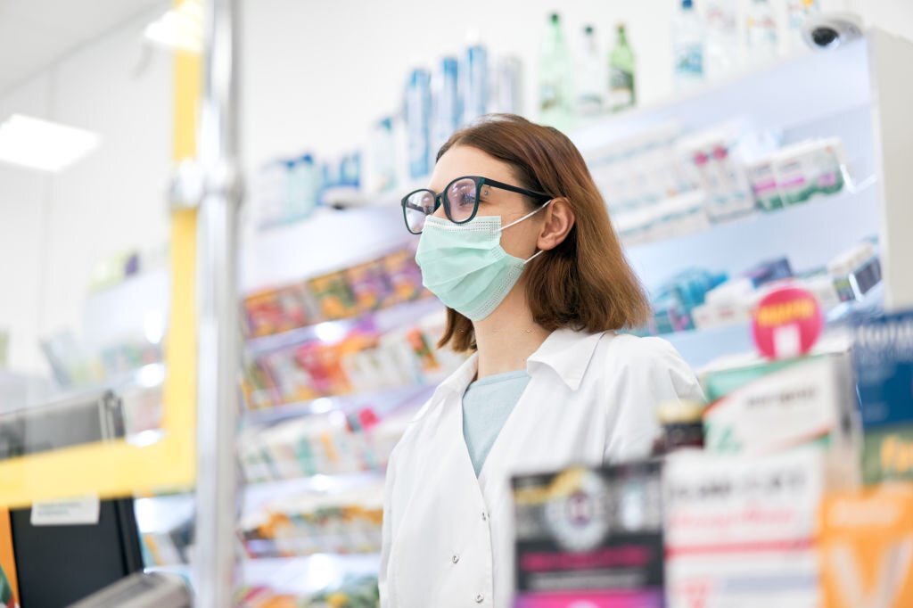Low angle of female pharmacist in face mask and glasses standing behind counter with cash register while working in pharmacy during epidemic.